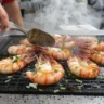 Langoustines en train de griller sur une plancha avec huile d’olive et persil, fumée légère, ambiance conviviale d’été, style photo culinaire appétissant.