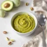 Purée pistache et avocat dans un petit bol, entourée de pistaches et d’avocat frais.